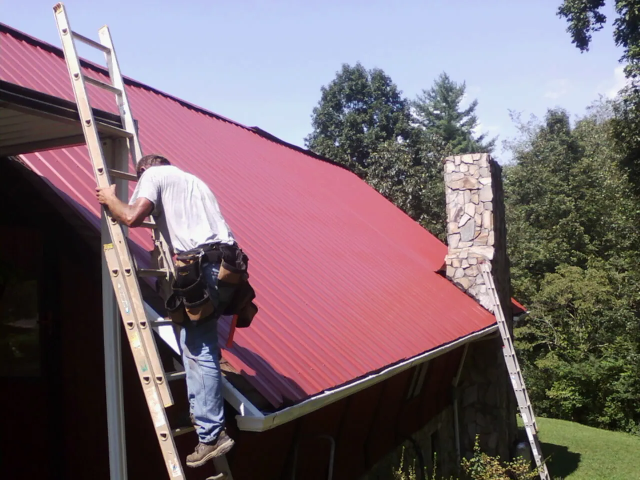 Roofer on a ladder beside a red metal roof and stone chimney — Soffit & Fascia Repair by Roofing Brothers in Pennsburg